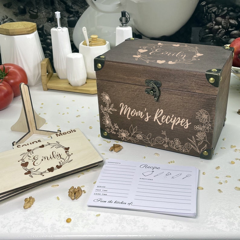 May include: A wooden recipe box with a floral design and the text "Mom's Recipes" and "Emily" engraved on the lid. The box is open and there are recipe cards inside. There is also a wooden recipe card holder with the text "Emily" engraved on it.