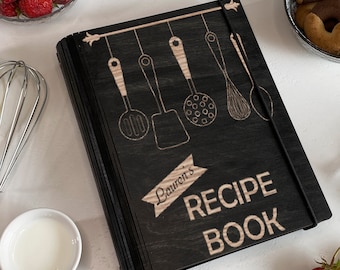 May include: Black wooden recipe book with a hanging kitchen utensil design and the text "Lauren's Recipe Book" in a light wood color.