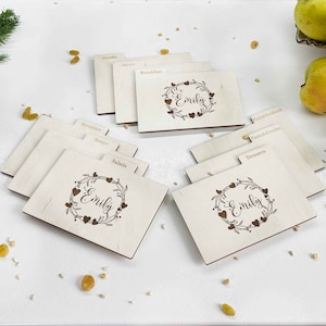 May include: Set of wooden recipe index cards with a floral wreath design and the name "Emily" engraved on each card. The cards are labeled with categories such as "Drinks", "Breakfast", "Salads", "Soups", "Favorites", "Fish & Seafood", "Pasta & Grains", "Desserts", and "Menu".