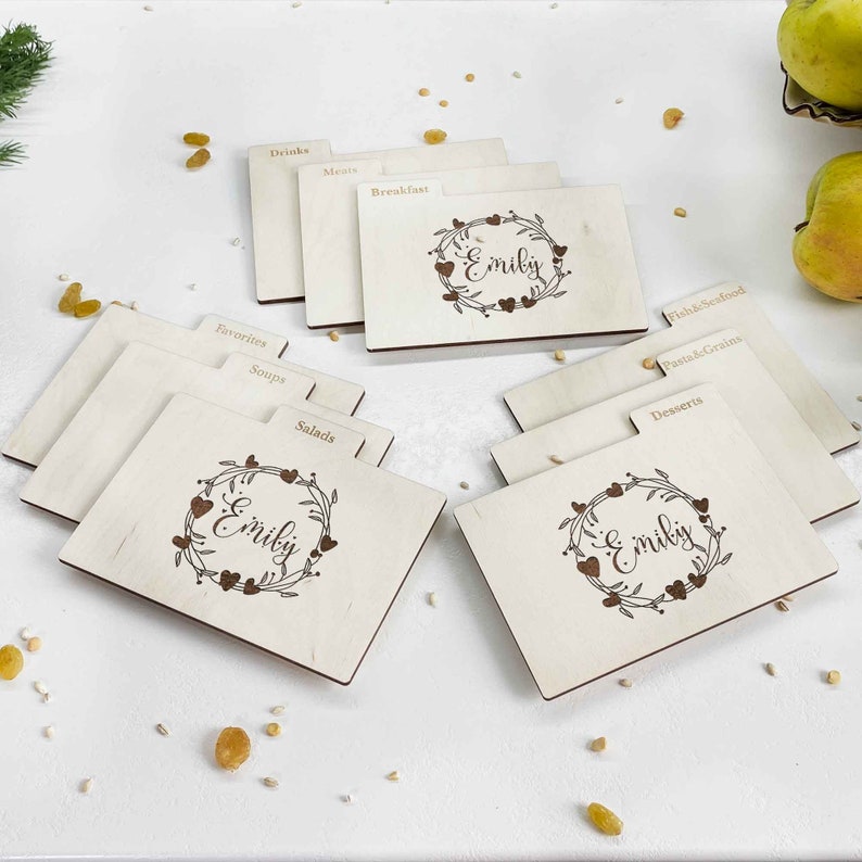 May include: Set of wooden recipe index cards with a floral wreath design and the name "Emily" engraved on each card. The cards are labeled with categories such as "Drinks", "Breakfast", "Salads", "Soups", "Favorites", "Fish & Seafood", "Pasta & Grains", "Desserts", and "Menu".