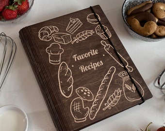 May include: A dark brown wooden recipe binder with a black elastic band closure. The cover is decorated with a laser-engraved design of various baking-themed illustrations, including bread, pastries, and a rolling pin. The text "Favorite Recipes" is also engraved on the cover.