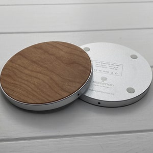 May include: A silver wireless charging pad with a wood veneer top. The pad has a label that reads "FAST WIRELESS CHARGER" and lists the input and output voltage and amperage. The label also includes the CE, FC, and RoHS certifications. The pad is made in Ukraine by SKINWOOD.