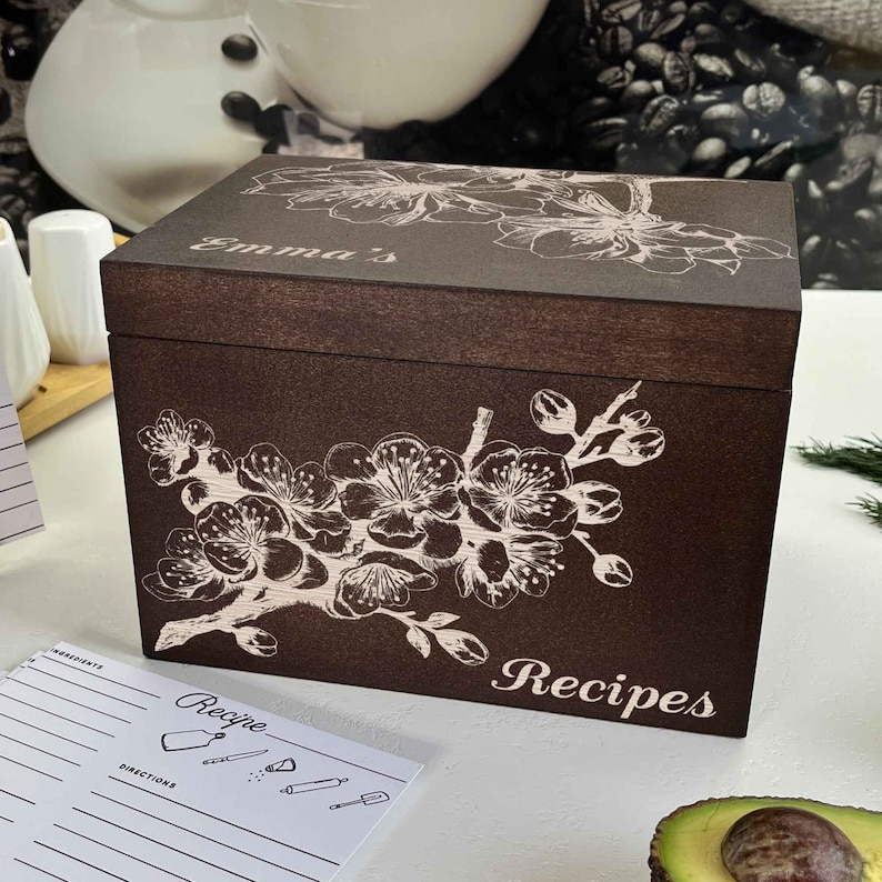 K&ouml;nnte beinhalten: Eine dunkelbraune Holz-Rezeptbox mit einem wei&szlig;en Blumenmuster und dem Text "Emma's Recipes".
