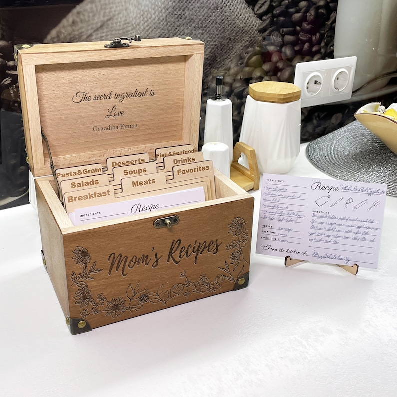 May include: A wooden recipe box with a hinged lid and a floral design. The box has a label that says "Mom's Recipes" and includes dividers for different recipe categories, such as "Pasta & Grain", "Desserts", "Soups", "Salads", "Breakfast", "Meats", "Fish & Seafood", "Drinks", and "Favorites". The box also has a small card that says "The secret ingredient is Love, Grandma Emma".