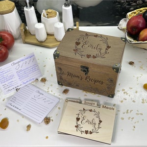 May include: A wooden recipe box with a floral design and the name "Emily" engraved on the lid. The box has a metal latch and is surrounded by recipe cards with dividers labeled "Desserts", "Soups", and "Meats".