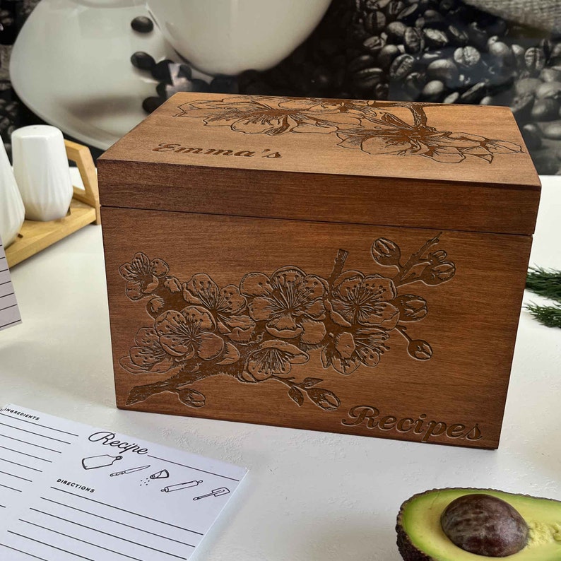 K&ouml;nnte beinhalten: Eine Holzkisten f&uuml;r Rezepte mit einem Blumendesign und dem Text "Emma's Recipes" auf der Vorderseite. Die Kiste ist geschlossen und steht auf einer wei&szlig;en Oberfl&auml;che.