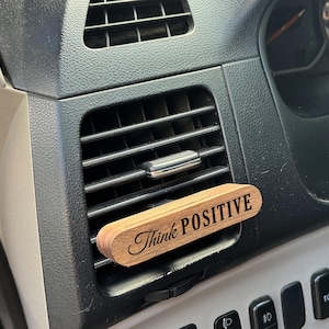 May include: A wooden car air freshener with the text "Think Positive" engraved on it. The air freshener is brown and has a natural wood grain. It is inserted into the air vent of a car.