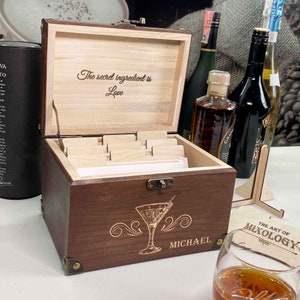 May include: A wooden box with a hinged lid, engraved with the text "The secret ingredient is Love". The box has a compartment for cards and is labelled "MICHAEL". The box is sitting on a table with a glass of brown liquid and a sign that reads "The Art of Mixology".
