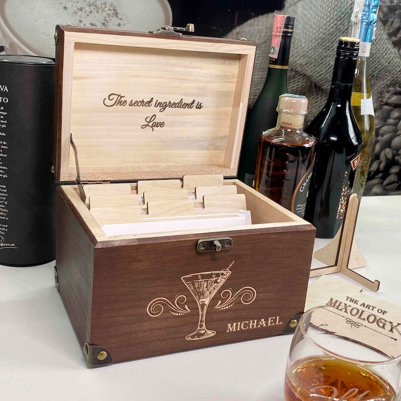 May include: A wooden box with a hinged lid, engraved with the text "The secret ingredient is Love". The box has a compartment for cards and is labelled "MICHAEL". The box is sitting on a table with a glass of brown liquid and a sign that reads "The Art of Mixology".