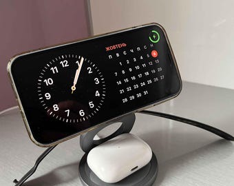 May include: A black smartphone with a clock face and calendar showing the month of October in Russian. The phone is on a black charging stand with a white wireless charging pad for earbuds.
