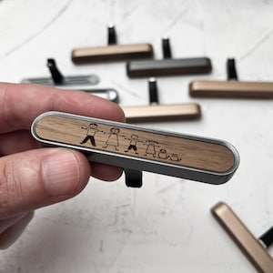 Può includere: Un profumatore per auto in argento con un top in legno impiallacciato. L'impiallacciatura presenta un design inciso a laser di una famiglia di figure stilizzate che si tengono per mano con un animale domestico.