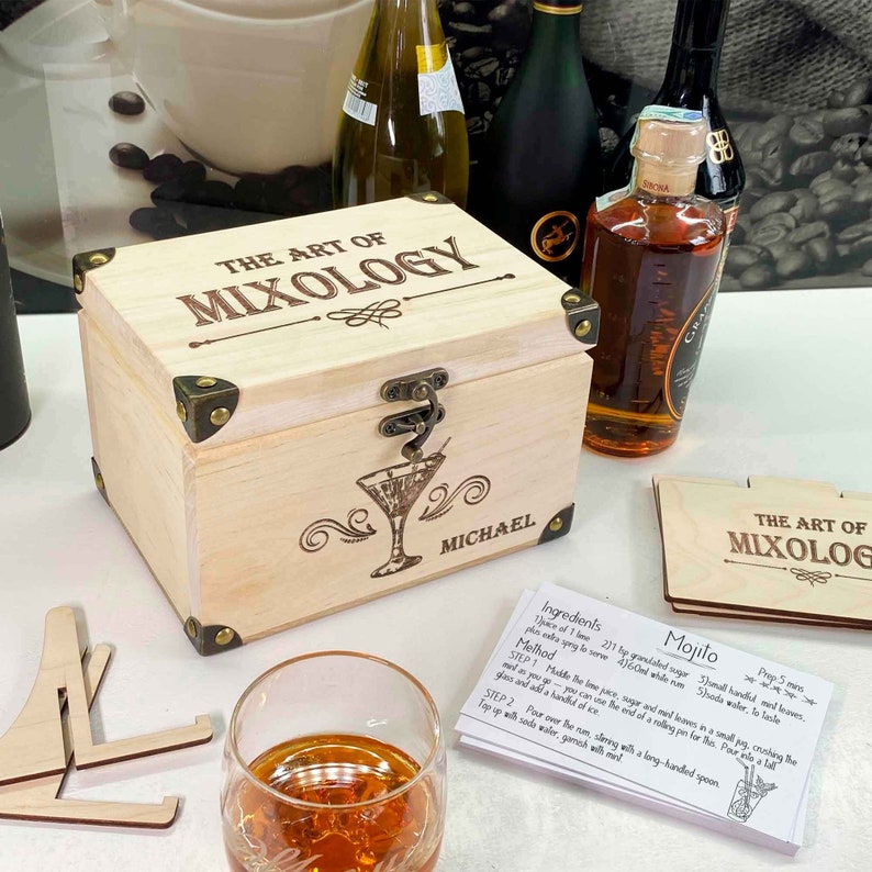 May include: A wooden box with the words "The Art of Mixology" engraved on the lid. The box is closed and has a brass latch. The box is personalised with the name "Michael". There are several cocktail recipe cards on the table next to the box.