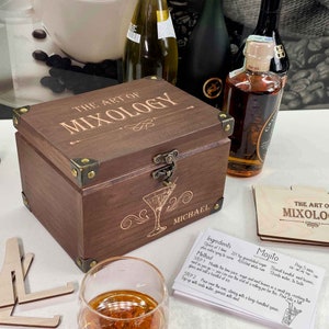 May include: A wooden box with the words "The Art of Mixology" engraved on the lid. The box is closed and has a brass latch. The box is sitting on a table with a glass of liquid and a card with the recipe for a Mojito.