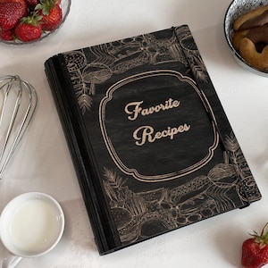 Può includere: Libro di ricette in legno nero con un'illustrazione in bianco e nero di cibo e il testo "Favorite Recipes" sulla copertina.