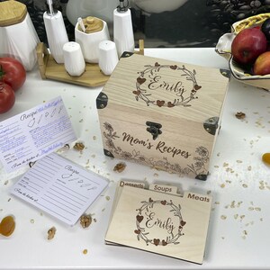 May include: Wooden recipe box with a floral design and the name "Emily" engraved on the front. The box has a hinged lid and a lock. The box is surrounded by recipe cards and other kitchen items.