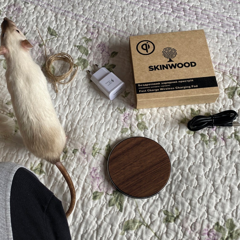 May include: A brown wooden wireless charging pad with a black trim and a white USB adapter. The charging pad is on a white and pink floral patterned surface. The text on the box reads "Skinwood Fast Charge Wireless Charging Pad".