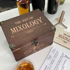 May include: A wooden box with a hinged lid and brass accents. The box is engraved with the text "The Art of Mixology" and "Michael". The box is open and a card with a recipe for a Mojito is visible.