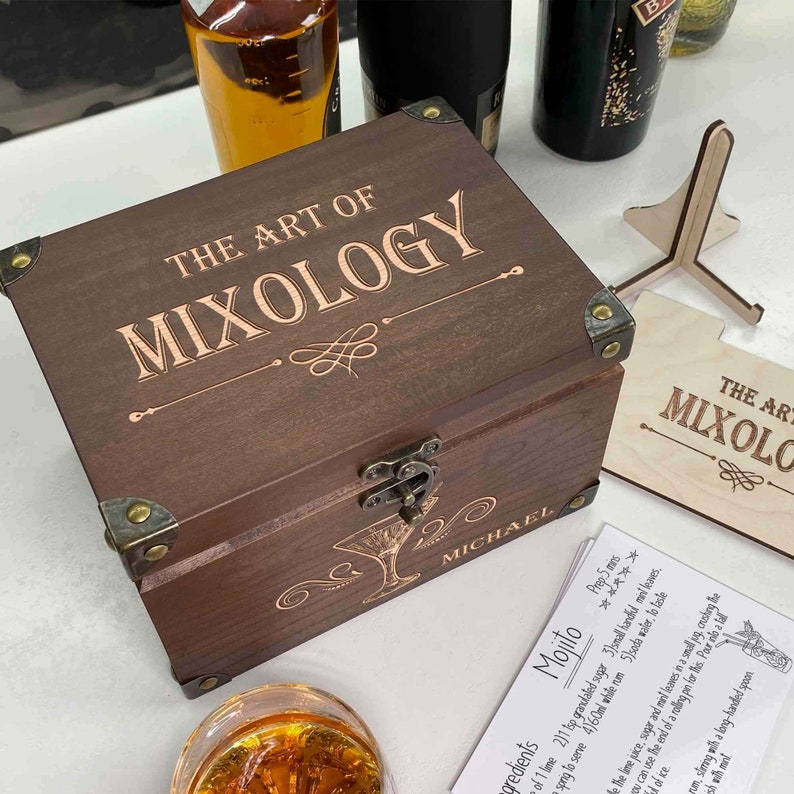 May include: A wooden box with a hinged lid and brass accents. The box is engraved with the text "The Art of Mixology" and "Michael". The box is open and a card with a recipe for a Mojito is visible.