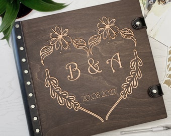 May include: A dark brown wooden photo album with a black leather spine and decorative stitching. The front cover features a heart-shaped design with floral accents and the initials "B&A" and the date "20.08.2022".