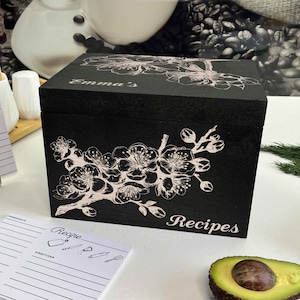 K&ouml;nnte beinhalten: Schwarze Rezeptbox aus Holz mit wei&szlig;em Blumendesign und dem Wort "Rezepte" auf der Vorderseite. Der Deckel tr&auml;gt den Namen "Emma's".