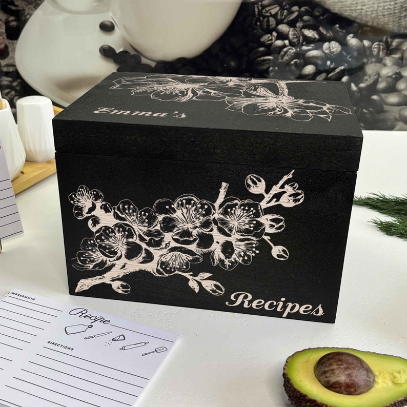K&ouml;nnte beinhalten: Schwarze Rezeptbox aus Holz mit wei&szlig;em Blumendesign und dem Wort "Rezepte" auf der Vorderseite. Der Deckel tr&auml;gt den Namen "Emma's".