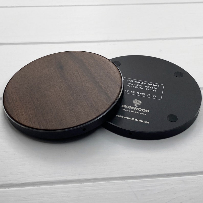 May include: A black wireless charging pad with a wood veneer top. The charging pad has a label on the bottom that reads "FAST WIRELESS CHARGER Input: 5V/2A 9V/1.67A Output: 5V/1A 9V/1.1A CE FC RoHS SKINWOOD Made in Ukraine skinwood.com.ua"