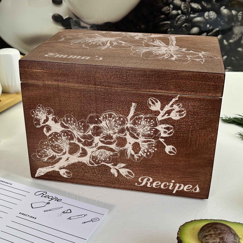 K&ouml;nnte beinhalten: Eine braune Holzkisten f&uuml;r Rezepte mit einem wei&szlig;en Blumendesign und dem Wort "Rezepte" auf der Vorderseite. Die Kiste ist mit einem wei&szlig;en Blumendesign verziert und der Name "Emma's" steht auf der Oberseite.