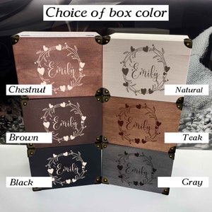 May include: Six wooden boxes with different wood finishes, each with a heart wreath design and the name "Emily" engraved on the lid. The boxes are labeled with the wood finishes: Chestnut, Natural, Teak, Brown, Gray, and Black.