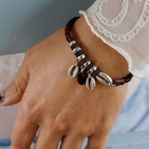 Könnte beinhalten: Ein braunes Lederarmband mit silbernen Muschel-Anhängern und Perlen. Das Armband befindet sich an einem Handgelenk.
