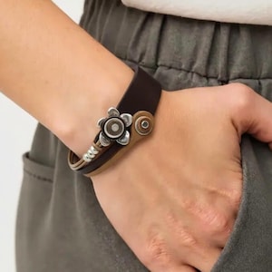 Könnte beinhalten: Ein braunes Lederarmband mit einem silbernen, blütenförmigen Verschluss. Das Armband hat ein zweifarbiges Design, mit einem dunkleren braunen Band oben und einem helleren braunen Band darunter. Der Verschluss hat ein kreisförmiges Design mit einer silbernen Blume.