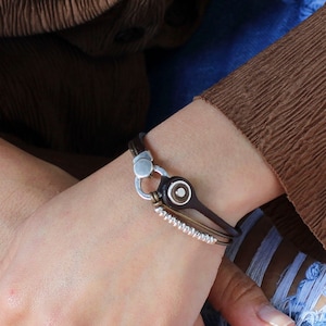 May include: A layered bracelet set featuring a brown leather bracelet with a silver clasp and a silver and brown charm, a silver chain bracelet with a silver bead accent, and a silver chain bracelet with a silver bead accent.