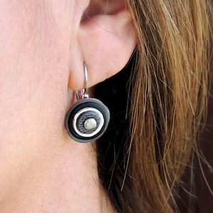 Puede incluir: Pendientes de plata con un colgante circular negro y plateado.