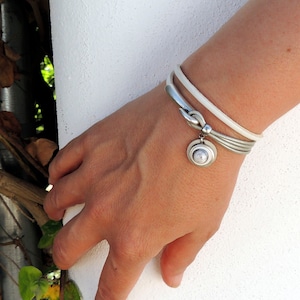 Peut inclure: Un bracelet en argent avec un bracelet en cuir blanc et un charm décoratif. Le bracelet comporte plusieurs bandes argentées et une fermeture à fermoir. Le charm a un design circulaire avec un centre blanc. Le bracelet est porté au poignet.