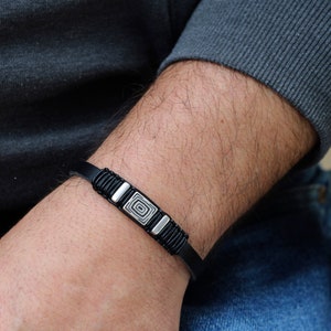 May include: A black leather bracelet with a silver square design. The bracelet is worn on a person's wrist.
