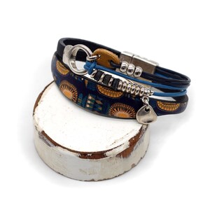 Könnte beinhalten: Mehrere Armbänder übereinander. Ein Armband in Marineblau mit gelbem Sonnenmuster, ein weiteres aus blauem Leder und ein drittes aus schwarzem Leder. Silberfarbene Details und ein Tropfenanhänger ergänzen das Design.