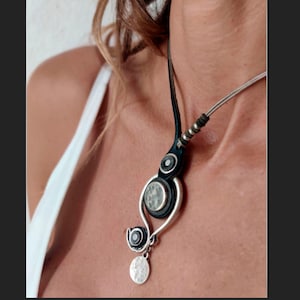 Pu&ograve; includere: Una collana argento e nera con un cordoncino in pelle e un ciondolo con un design circolare con una pietra bianca. Il ciondolo &egrave; appeso a una catena d'argento con un ciondolo circolare.