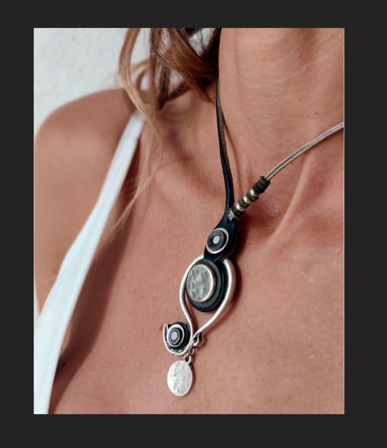 Pu&ograve; includere: Una collana argento e nera con un cordoncino in pelle e un ciondolo con un design circolare con una pietra bianca. Il ciondolo &egrave; appeso a una catena d'argento con un ciondolo circolare.