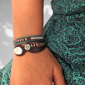 Könnte beinhalten: Ein mehrlagiges Armband aus braunem Leder, silbernen Perlen und türkisfarbenen Akzenten. Das Armband wird am Handgelenk getragen.