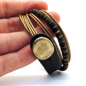 May include: A multi-strand wrap bracelet with gold-coloured cords, black leather accents, and a brass-coloured button clasp. The design includes a section of small, dark beads. This bracelet is a stylish accessory, suitable for everyday wear.