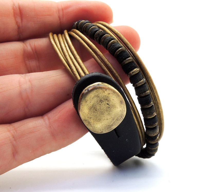 May include: A multi-strand wrap bracelet with gold-coloured cords, black leather accents, and a brass-coloured button clasp. The design includes a section of small, dark beads. This bracelet is a stylish accessory, suitable for everyday wear.