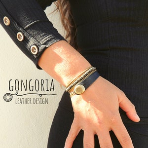 May include: A wrist featuring a stack of gold-coloured bangles and a dark blue leather bracelet with a large, round, gold-coloured button clasp. The image also shows the text "GONGORIA LEATHER DESIGN".