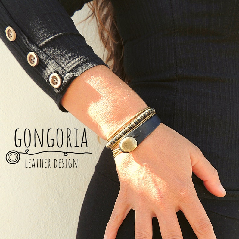May include: A wrist featuring a stack of gold-coloured bangles and a dark blue leather bracelet with a large, round, gold-coloured button clasp. The image also shows the text "GONGORIA LEATHER DESIGN".