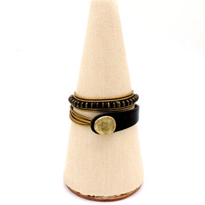 May include: A selection of rings presented on a beige, cone-shaped display stand. The rings feature a black leather band with a gold-coloured button, thin gold-coloured bands, and a dark bronze-coloured ring with a textured pattern. The background is white.