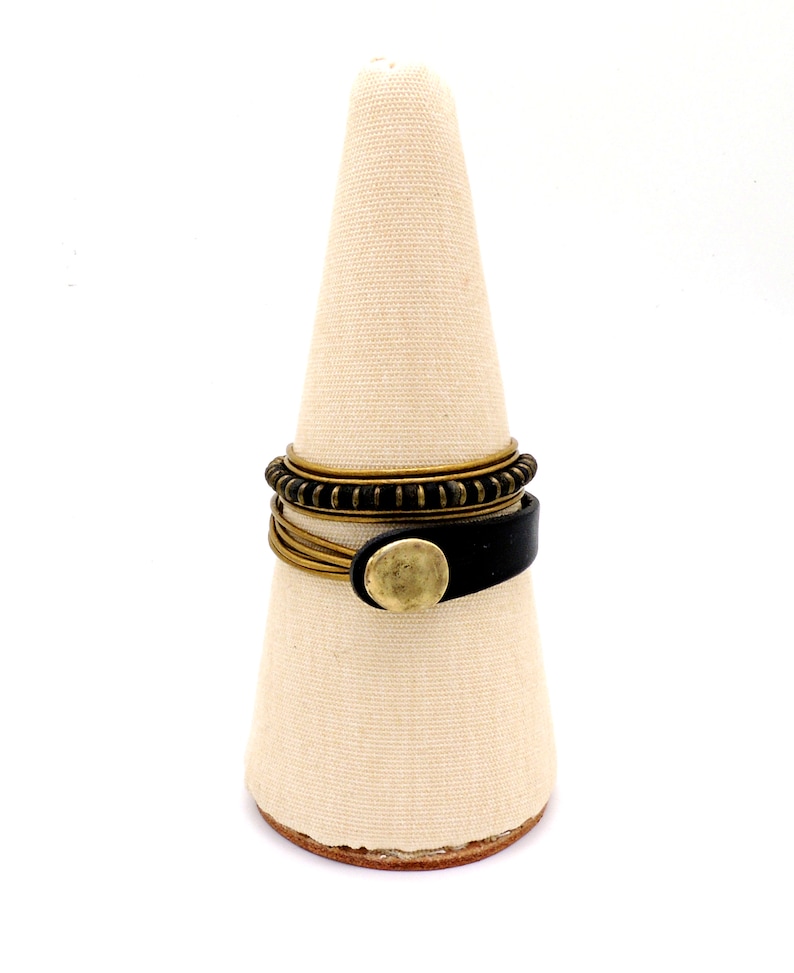 May include: A selection of rings presented on a beige, cone-shaped display stand. The rings feature a black leather band with a gold-coloured button, thin gold-coloured bands, and a dark bronze-coloured ring with a textured pattern. The background is white.