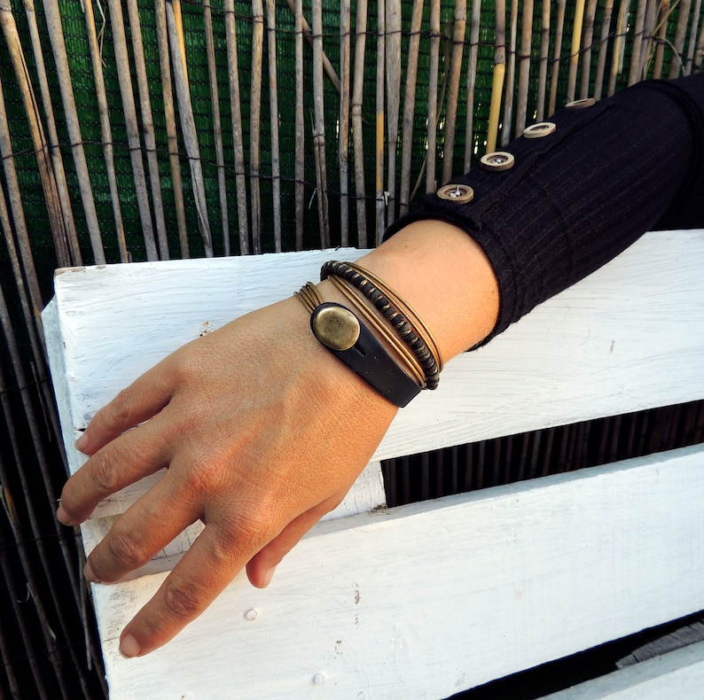 May include: A hand adorned with multiple bracelets. One bracelet is black leather with a gold-coloured oval accent. Other bracelets are gold-coloured metal and beaded. The person wears a black long-sleeved shirt with gold-coloured buttons.