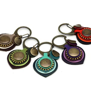 Puede incluir: Cinco llaveros de cuero coloridos con anillos y dijes de metal. Cada llavero presenta un diseño en capas con un dije circular de metal en el centro. Los llaveros son en tonos de marrón, verde, morado, azul y rojo.