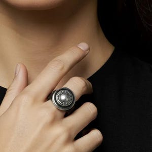 Puede incluir: Anillo plateado con diseño circular. El anillo presenta una esfera central elevada rodeada por un anillo de esferas más pequeñas. El anillo se lleva en un dedo, con fondo negro.