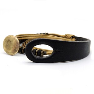 May include: A leather bracelet with a black strap and a gold-coloured, circular clasp. The bracelet features multiple strands of tan-coloured leather and small, dark beads. The design is simple and rustic, suitable for everyday wear.
