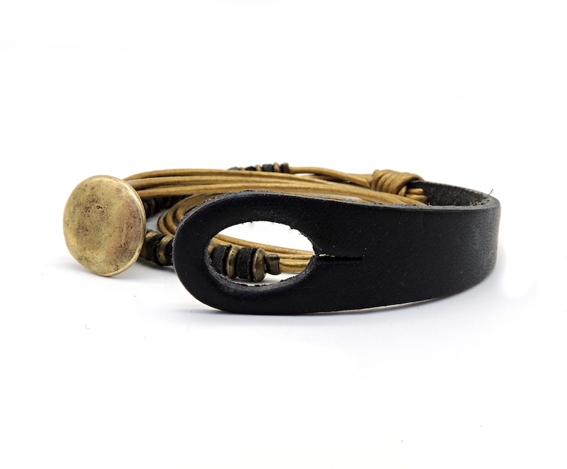 May include: A leather bracelet with a black strap and a gold-coloured, circular clasp. The bracelet features multiple strands of tan-coloured leather and small, dark beads. The design is simple and rustic, suitable for everyday wear.
