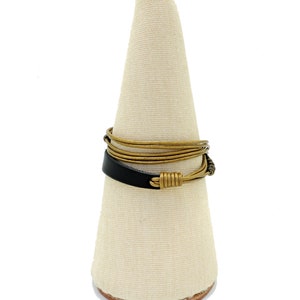 May include: A handcrafted ring with a black leather band and gold-toned wire detailing. The ring is displayed on a white cone-shaped stand, highlighting its unique design. The ring's design incorporates a mix of materials and textures, showcasing a modern aesthetic.